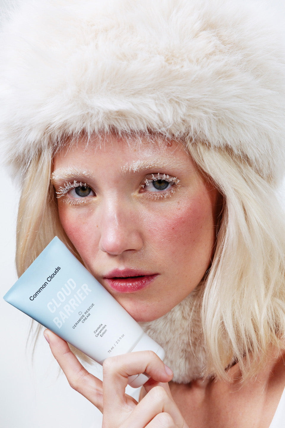 Person wearing a white fur hat holding a Common Clouds Cloud Barrier skincare product with a plain background.
