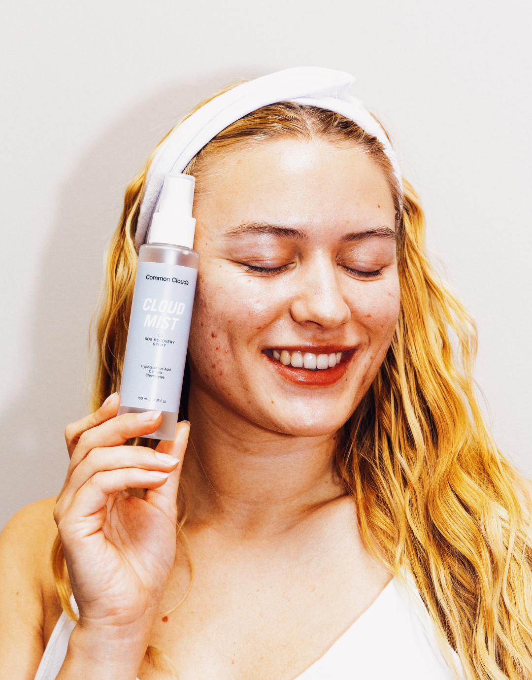 Smiling woman with acne-prone skin and long, wavy blonde hair holds a bottle of Common Clouds "Cloud Mist" SOS Recovery Spray next to her face.