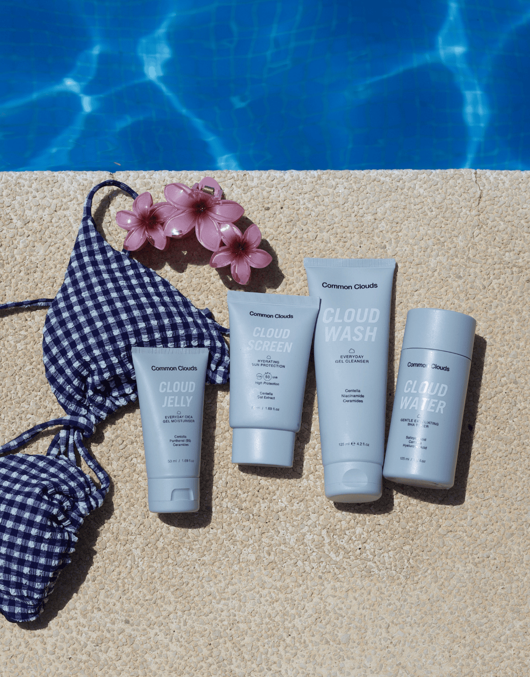 Common Clouds skincare products by a pool with a bikini top and flower clip.