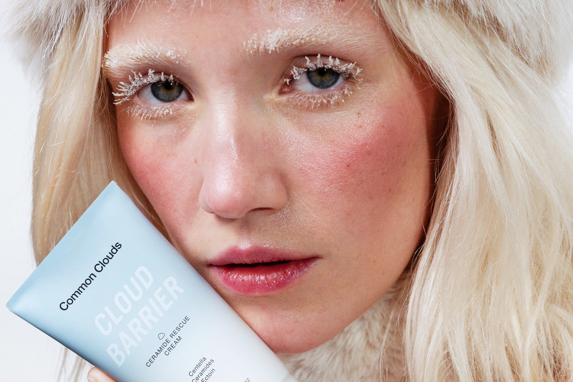 Person wearing a white fur hat holding a skincare product against a white background