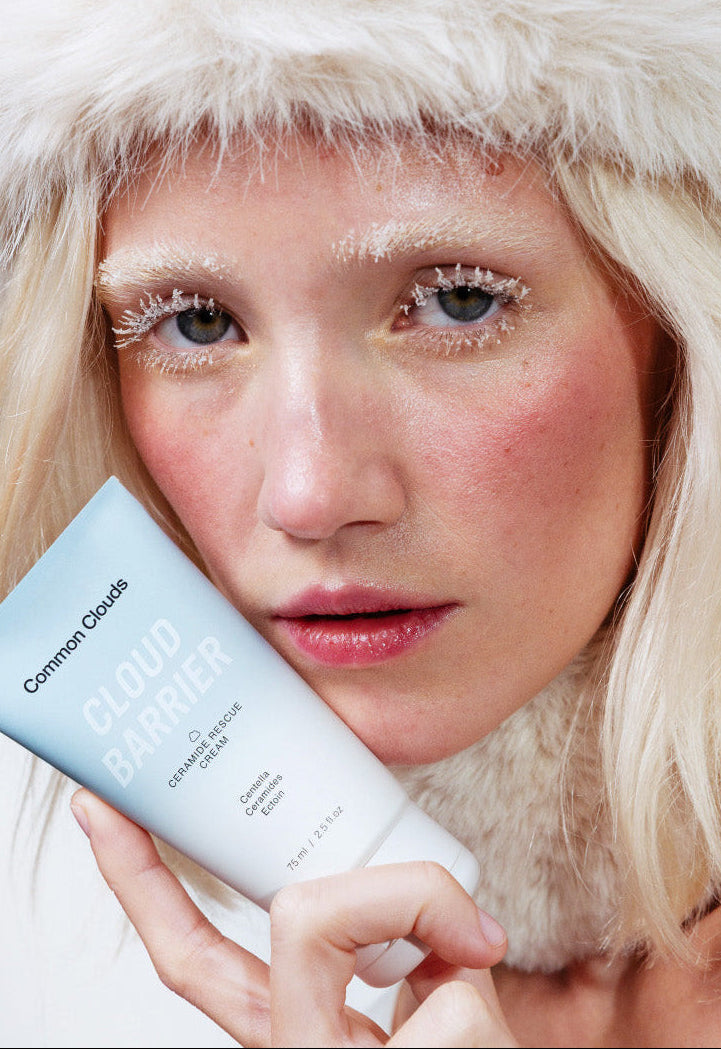 Person wearing a white fur hat holding a skincare product with a plain background