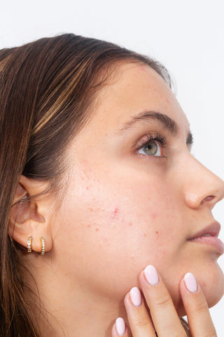 Close-up side profile of a woman with clear skin and small gold hoop earrings, touching her cheek against a plain light background.