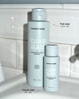 Two Common Clouds Cloud Glaze Hydrating Barrier Essence bottles on a white tiled bathroom counter, showing the full size 125 ml and travel size 40 ml.