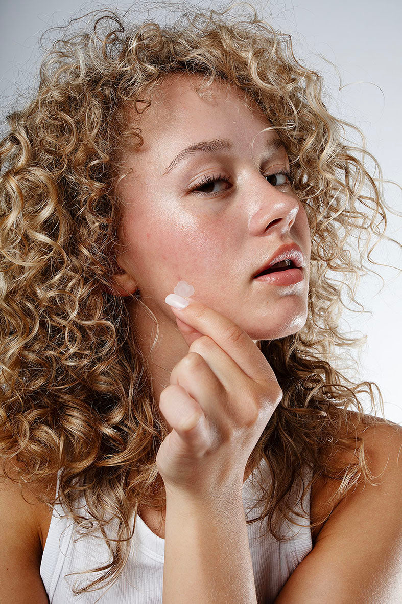 Woman with curly blonde hair applying a Common Clouds Cloud Patch to her cheek, wearing a white tank top against a neutral background.