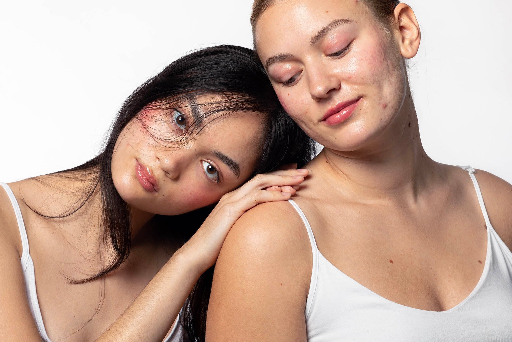 Two women wearing white tank tops against a plain background