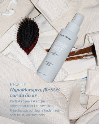A bottle of Clouds Cloud Spray lies on a beige fabric surface with a black hairbrush, white earbuds, and other small styling props.